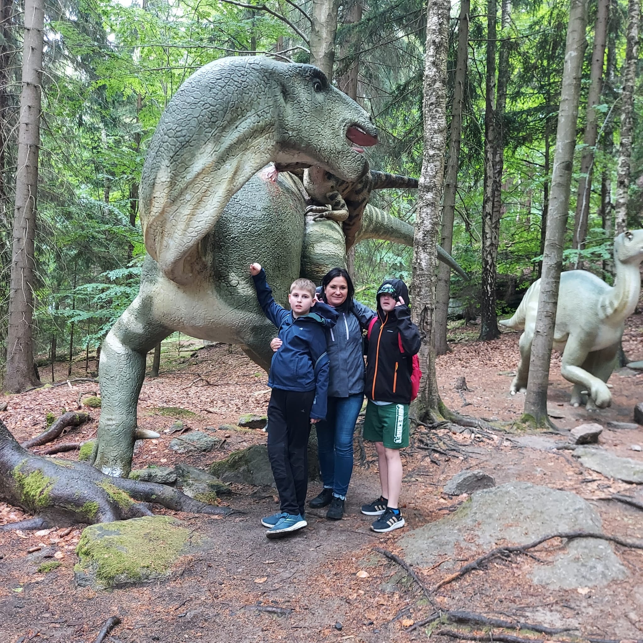 Grupa osób stoi przed modelem dinozaura w lesie 
