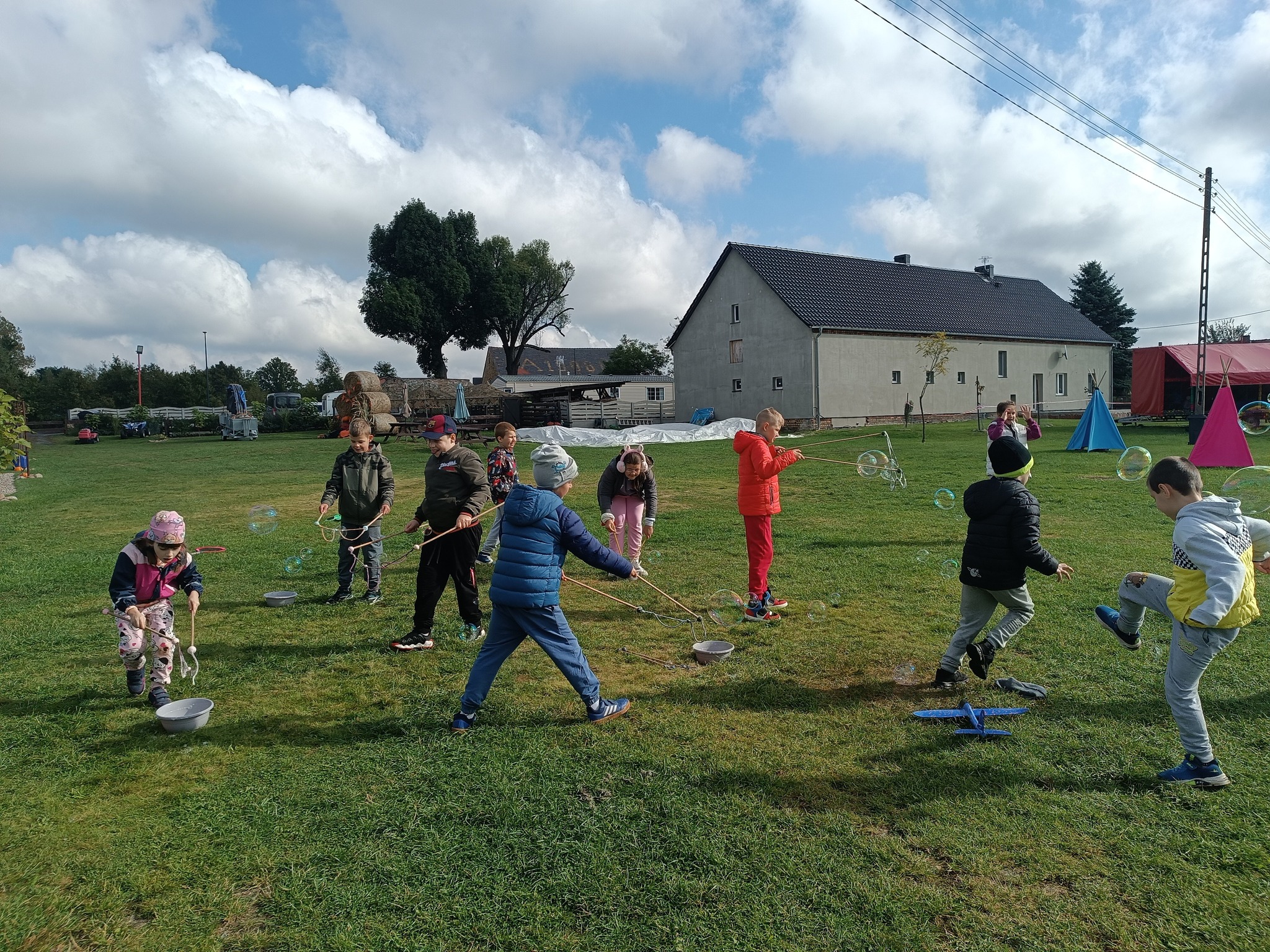 Na obrazku widać: grupę osób, dom, drzewo, niebo, trawę