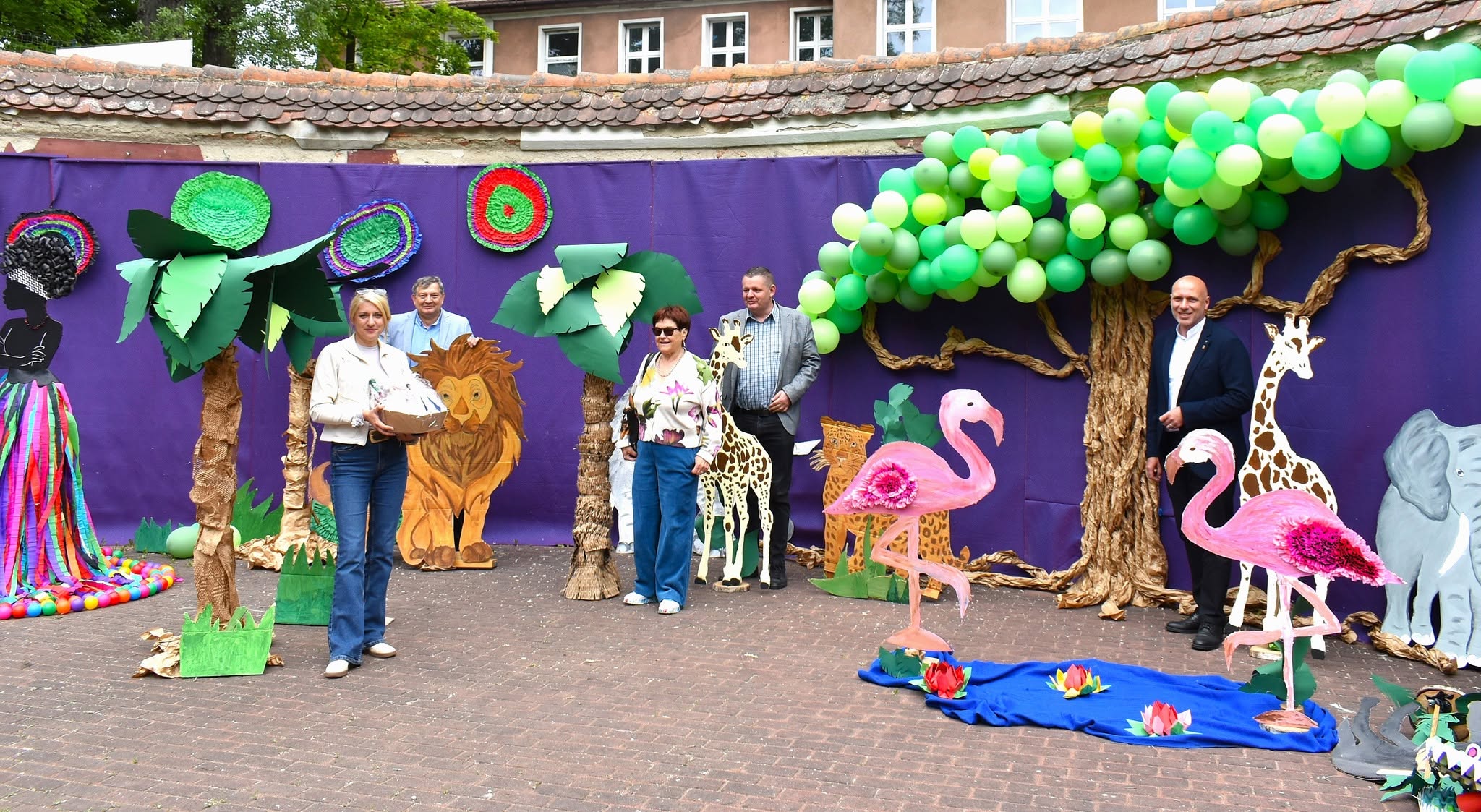 Na obrazku widać: grupę osób, makiety zwierzat, drzew, balony.